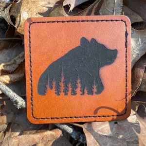 May include: A brown leather coaster with a black silhouette of a bear standing in a forest. The bear is facing the right side of the image.