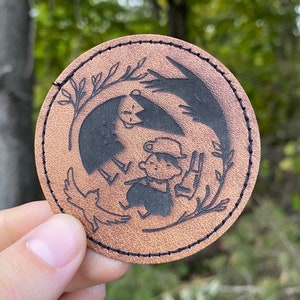 May include: A brown leather patch with a black and white illustration of a person with a bird and a tree. The patch is stitched around the edges.