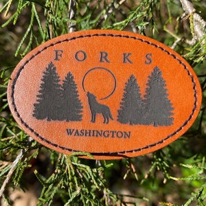 May include: A brown leather oval-shaped tag with black stitching. The tag has the text "FORKS WASHINGTON" and a silhouette of a wolf howling at the moon with three pine trees in the background.