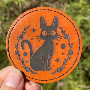 May include: A round, orange leather coaster with a black cat silhouette and floral design. The cat has large, round eyes and a whimsical expression.