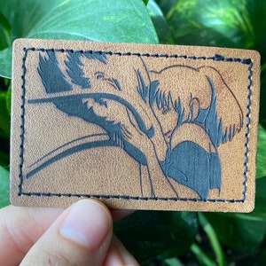 May include: A brown leather patch with black stitching around the edges. The patch features a black silhouette of two figures embracing.