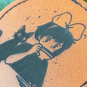 May include: A brown leather coaster with a black silhouette of a girl with a bow in her hair and a black cat. The girl is holding a cup and the cat is sitting next to her.