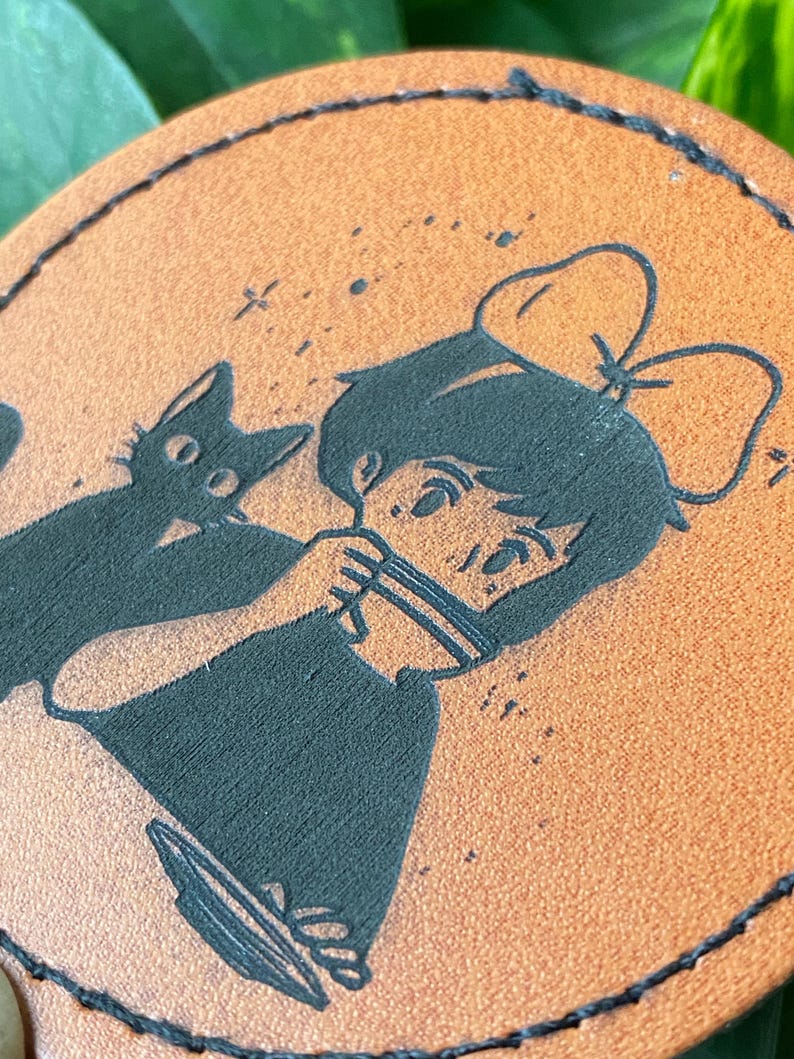 May include: A brown leather coaster with a black silhouette of a girl with a bow in her hair and a black cat. The girl is holding a cup and the cat is sitting next to her.