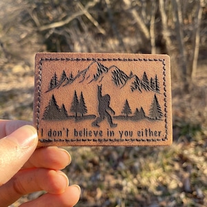 May include: A brown leather patch with a black silhouette of a Bigfoot standing in a forest with mountains in the background. The text "I don't believe in you either" is printed below the image.