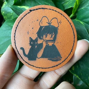 May include: A brown leather coaster with a black silhouette of a girl with a bow in her hair drinking from a cup and a black cat sitting next to her.
