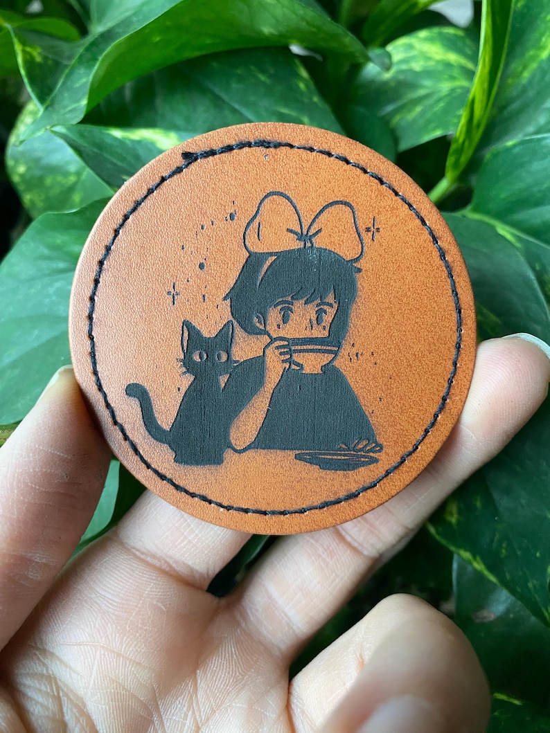 May include: A brown leather coaster with a black silhouette of a girl with a bow in her hair drinking from a cup and a black cat sitting next to her.