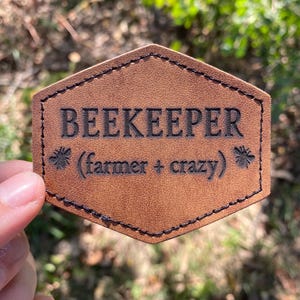 May include: A brown leather patch with black stitching and the text "BEEKEEPER (farmer + crazy)" with a bee icon on either side of the text.