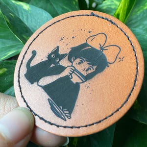 May include: A brown leather coaster with a black and white illustration of a girl with a black cat. The girl is wearing a bow and has her hair pulled back. The cat is sitting on her shoulder.