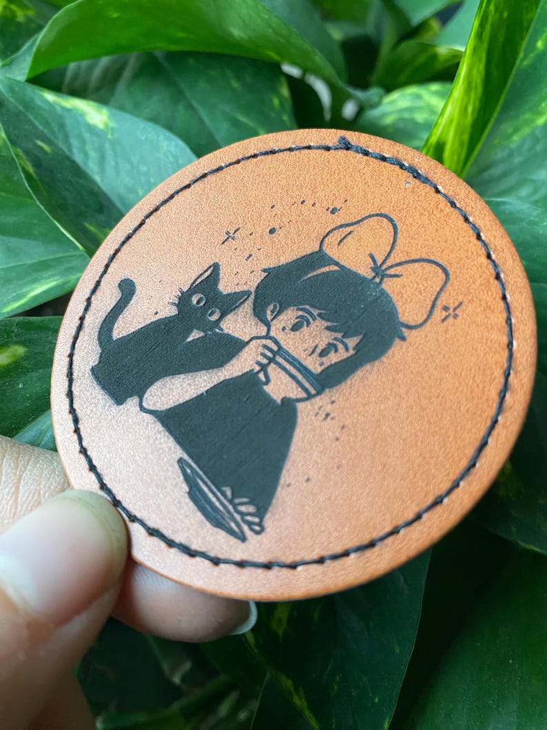 May include: A brown leather coaster with a black and white illustration of a girl with a black cat. The girl is wearing a bow and has her hair pulled back. The cat is sitting on her shoulder.