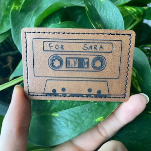 May include: A brown leather cassette tape keychain with black stitching and the words "For Sara" written on it.