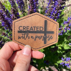 May include: A brown leather hexagon-shaped tag with the text "CREATED with a purpose" and a black cross.