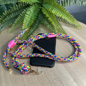 Può includere: Un cordino per telefono arcobaleno con una custodia nera. Il cordino è realizzato in corda intrecciata in una varietà di colori, tra cui rosa, giallo, blu e verde. Il cordino ha dettagli metallici color oro e una chiusura.