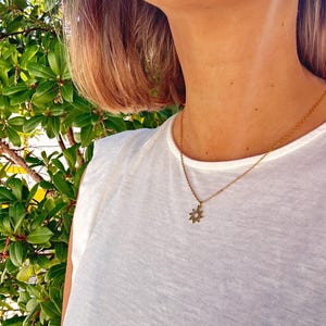 Op de afbeelding: Een goudkleurige ketting met een zonnestraal hanger. De hanger heeft een gedetailleerd ontwerp met kleine, delicate kenmerken. De ketting wordt gedragen over een wit t-shirt, met een groene, bladerrijke achtergrond.