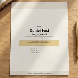 May include: A white 21-Day Daniel Fast Prayer Journal with gold text and a gold pen. The journal includes the text "Surrender - Seek - Receive" and a quote. The journal is open on a wooden surface.