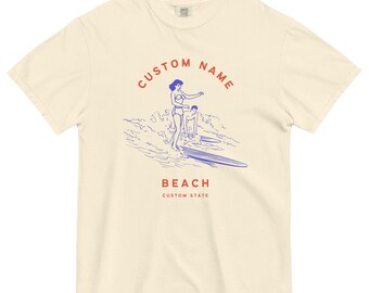 Puede incluir: Una camiseta de color crema con un gráfico azul y rojo de una mujer surfeando. El texto "CUSTOM NAME" está por encima del gráfico y "BEACH CUSTOM STATE" está por debajo del gráfico.