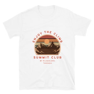 Puede incluir: Camiseta blanca con un gráfico retro de montañas y una puesta de sol. El gráfico dice "Enjoy the Climb" y "Summit Club" con el texto "Mt Kilimanjaro, Tanzania" debajo.