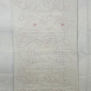 Op de afbeelding: Een wit stoffen paneel met een herhalend patroon van rode geborduurde ornamenten. De ornamenten zijn gestileerd en bevatten sterren, cirkels en gebogen lijnen.