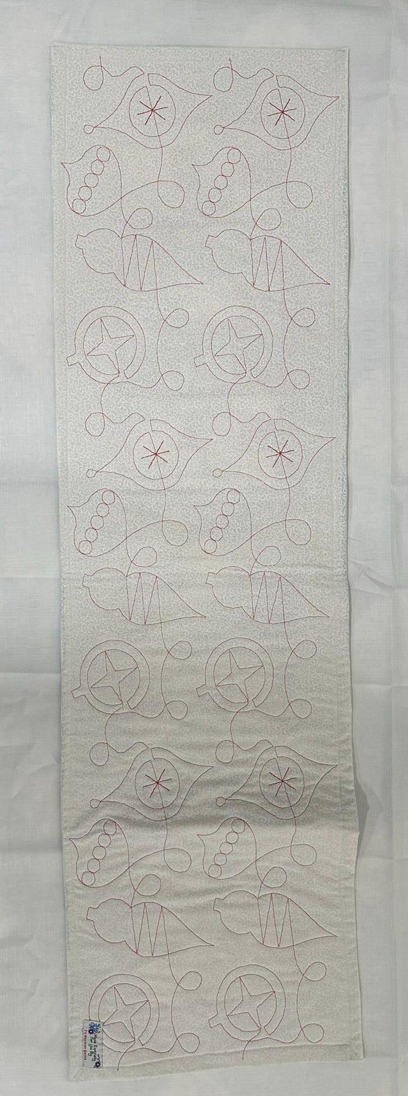 Op de afbeelding: Een wit stoffen paneel met een herhalend patroon van rode geborduurde ornamenten. De ornamenten zijn gestileerd en bevatten sterren, cirkels en gebogen lijnen.