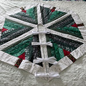 May include: A hexagonal Christmas tree skirt featuring a patchwork design of dark green, forest green, and black fabric strips with small red accents. The border is white fabric with a gray swirl pattern, secured by four white ribbon ties.