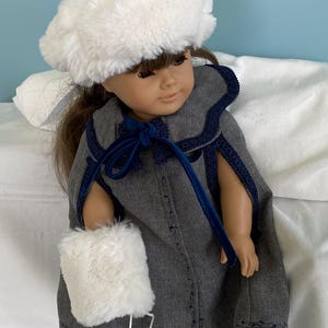May include: A doll dressed in a grey cape with navy blue trim, a white faux fur hat and muff. The cape features a navy blue velvet bow and decorative stitching. The doll is on a white surface.