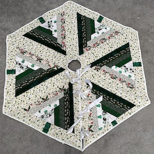 May include: A quilted Christmas tree skirt with a white floral pattern and green geometric accents. The skirt has a white border and is tied with white ribbons. The design includes various shades of green and white, creating a festive holiday look.
