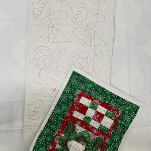Op de afbeelding: Een witte stoffen tafelloper met een rood en groen gestikt ontwerp. De loper heeft een sneeuwvlokkenpatroon en een rood en groen geruit ontwerp. De loper is dubbelgevouwen en ligt op een wit oppervlak.