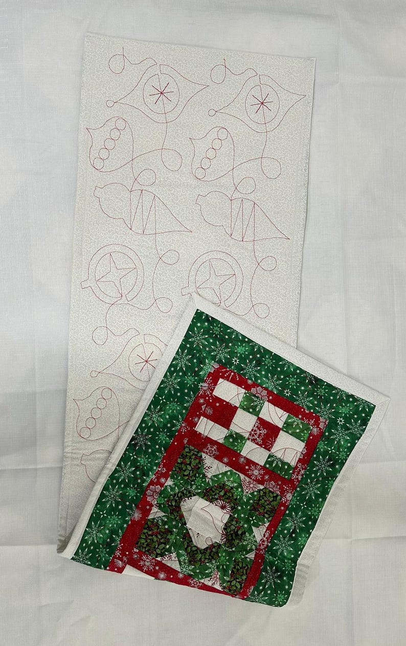 Op de afbeelding: Een witte stoffen tafelloper met een rood en groen gestikt ontwerp. De loper heeft een sneeuwvlokkenpatroon en een rood en groen geruit ontwerp. De loper is dubbelgevouwen en ligt op een wit oppervlak.