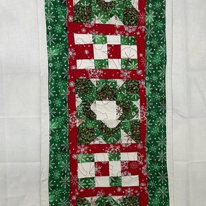 Könnte beinhalten: Ein grüner und roter gesteppter Tischläufer mit einem Schneeflockenmuster. Der Läufer hat einen weißen Rand und ist aus Stoff mit einem gesteppten Design.