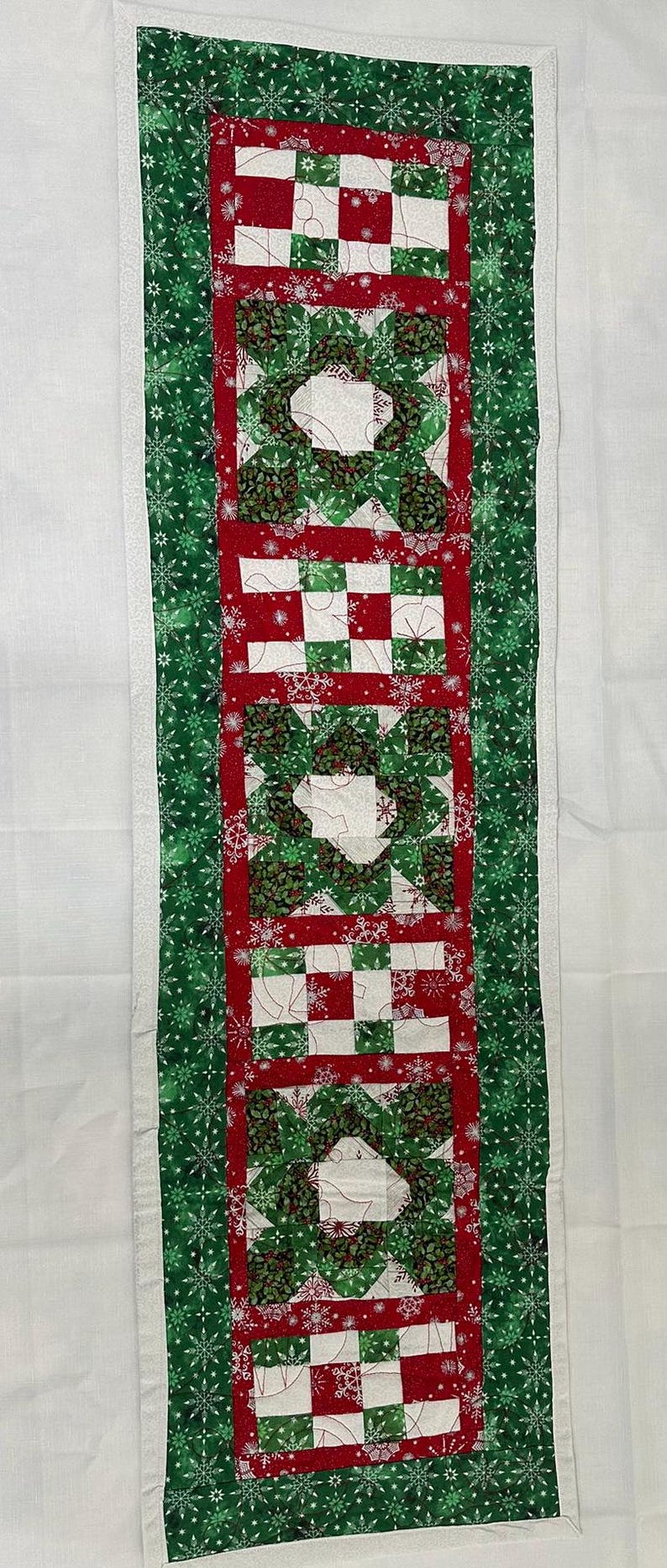 Op de afbeelding: Een groene en rode gequilte tafelloper met een sneeuwvlokkenpatroon. De loper heeft een witte rand en is gemaakt van stof met een gequilt ontwerp.