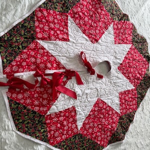 May include: A quilted Christmas tree skirt featuring a central white star with intricate stitching, surrounded by red fabric panels with white snowflake patterns and dark green fabric panels with red holly berry and leaf prints, finished with red ribbon ties.