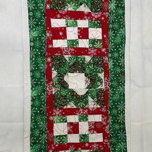 Op de afbeelding: Een groene en rode kersttafelrunner met een sneeuwvlokkenpatroonrand en een patchworkontwerp in het midden.