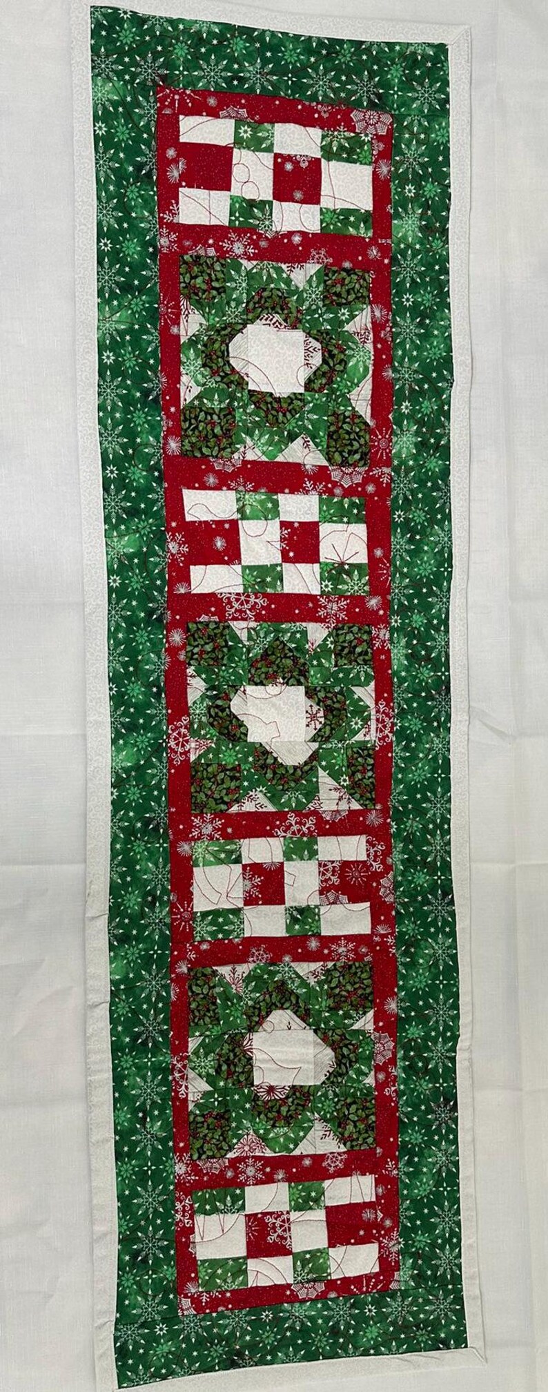 Op de afbeelding: Een groene en rode kersttafelrunner met een sneeuwvlokkenpatroonrand en een patchworkontwerp in het midden.