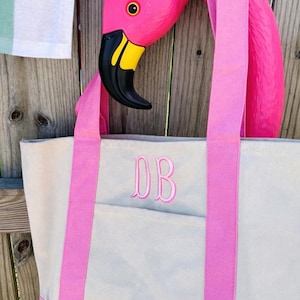 May include: A pink flamingo head decorates a canvas tote bag with pink straps and trim. The bag has a light gray body and a front pocket. The initials "DB" are embroidered in pink on the front of the bag. The flamingo has a black beak with yellow accents.