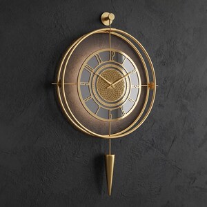 Könnte beinhalten: Eine goldfarbene Wanduhr mit einem kreisförmigen Rahmen aus konzentrischen Ringen, einem dunkelgrauen Zifferblatt mit goldenen römischen Ziffern und einem zentralen geometrischen Muster. Sie hat dünne goldene Zeiger und ein spitzes Pendel.