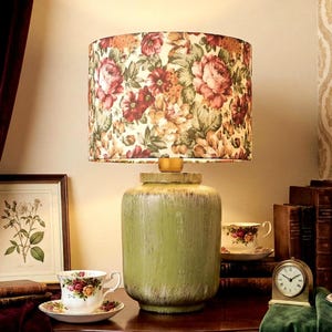 May include: A table lamp with a cylindrical green ceramic base and a drum lampshade featuring a colourful floral pattern of red, pink, and yellow roses on a cream background. The lamp sits on a dark wooden surface alongside two teacups.
