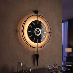 May include: A circular wall clock featuring a black face with white numerals and hands, surrounded by a thin black metal frame. The clock includes a downward-pointing black pendulum and emits a warm, circular glow from behind the clock face.