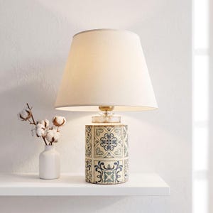Può includere: Una lampada da tavolo con un paralume conico in tessuto color crema e una base cilindrica decorata con un motivo di piastrelle blu e bianche. Un piccolo vaso bianco contenente steli di cotone essiccato si trova accanto alla lampada.