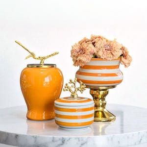 Könnte beinhalten: Drei dekorative Keramikteile: eine orangefarbene Vase mit goldenem Astdeckel, ein kleines gestreiftes Glas mit goldenem Korallen Deckel und eine gestreifte Vase mit Blumen. Die Vasen sind orange und weiß mit goldenen Akzenten.