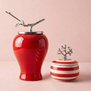 May include: Two decorative ceramic jars. The larger jar is red with a silver branch-shaped lid handle. The smaller jar is red and white striped with a silver coral-shaped lid handle. Both jars are on a pink surface.