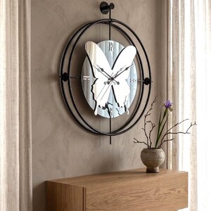 Op de afbeelding: Een ronde wandklok met een zwart metalen frame en een centrale spiegelende vlindervorm. De wijzerplaat bevat zwarte wijzers en zilverkleurige cijfers op de posities 12, 3, 6 en 9 uur, gemonteerd op een verticale zwarte metalen stang.