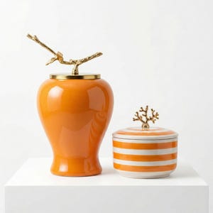 May include: Two decorative ceramic jars. The larger jar is orange with a gold branch-shaped handle. The smaller jar is white with orange horizontal stripes and a gold coral-shaped handle. Both jars are on a white surface.