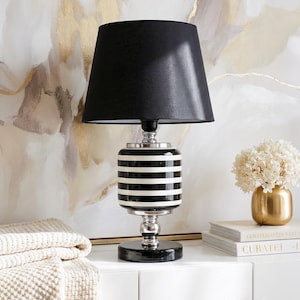 May include: A stylish table lamp featuring a black lampshade and a base with black and white stripes. The lamp has a chrome base and sits on a black circular base. A gold vase with white flowers is also visible.