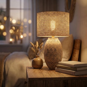 Puede incluir: Una lámpara de mesa beige con una pantalla texturizada y una base estampada se encuentra sobre una mesita de noche de madera. Un pequeño jarrón con flores secas y una pila de libros también están sobre la mesa. La lámpara está encendida.