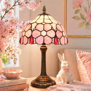 May include: A decorative table lamp with a stained glass shade featuring pink and red floral designs. The lamp has a bronze-coloured base and stem. A white ceramic bunny figurine with a pink bow sits nearby, along with a pink bowl and books.