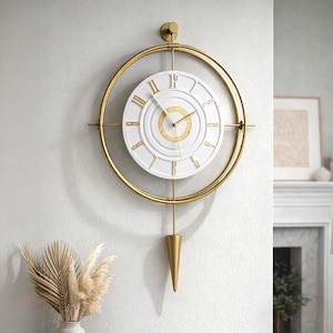 Puede incluir: Un reloj de pared de metal dorado con esfera blanca y números romanos. El reloj está rodeado por un marco circular dorado con un péndulo. El reloj está montado en una pared blanca. El diámetro del reloj es de aproximadamente 30 cm.