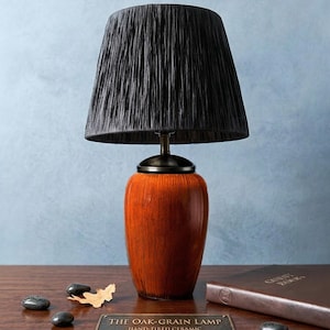 Puede incluir: Una lámpara de mesa decorativa con una pantalla negra y una base de cerámica con acabado de veta de roble. La lámpara tiene un cuello color bronce y una base que dice "THE OAK-GRAIN LAMP HAND-FIRED CERAMIC ETSY EXCLUSIVE."