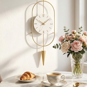 Pode incluir: Um relógio de parede dourado com mostrador branco e algarismos e ponteiros dourados. O relógio está suspenso dentro de uma moldura oval dourada e tem um pêndulo dourado. Um croissant, café e flores estão em uma mesa.