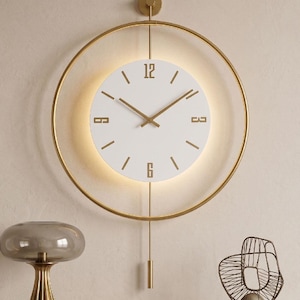 Könnte beinhalten: Eine kreisförmige Wanduhr mit weißem Zifferblatt und goldenen Ziffern auf 12, 3, 6 und 9 Uhr, umgeben von einem dünnen goldenen Metallring. Die Uhr verfügt über goldene Zeiger, eine vertikale Stange mit Pendelgewicht und eine integrierte Hintergrundbeleuchtung.