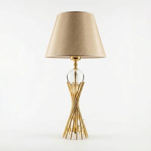 Puede incluir: Una lámpara de mesa dorada con una pantalla beige. La lámpara presenta un globo de cristal transparente y una base torcida y geométrica. La pantalla tiene una forma cónica clásica. La lámpara está diseñada para uso en interiores.