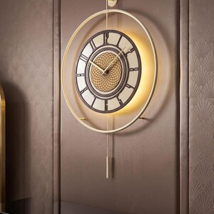 May include: A circular wall clock featuring a gold-toned frame, a dark brown dial with Roman numerals, and a central gold geometric grid pattern. The clock includes gold hands, a vertical pendulum, and integrated backlighting behind the dial.